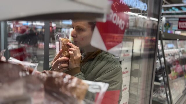 Woman inspects packaged meat at supermarket counter, turning clear wrapped product in her hands. Grocery shopper studies label with focused attention while choosing food from refrigerated display.