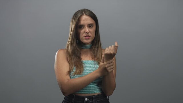 Woman holding and rubbing her wrist with a pained frown while checking the joint, wearing a turquoise top, choker and jeans against a grey studio backdrop; injury concern.