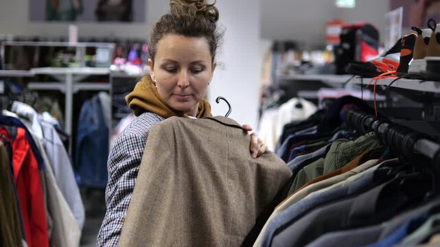 Woman examines brown jacket while shopping for clothing, lifting it from store rack and checking its fabric and fit. Thoughtful shopper compares garment details before making consumer choice.