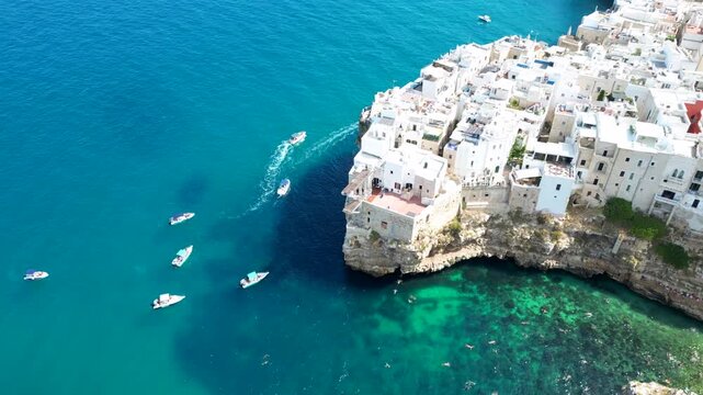 Aerial view of white buildings perched on limestone cliffs overlooking the turquoise sea with small boats navigating the coastal waters and Lama Monachile beach in Polignano a Mare, Apulia, Italy.