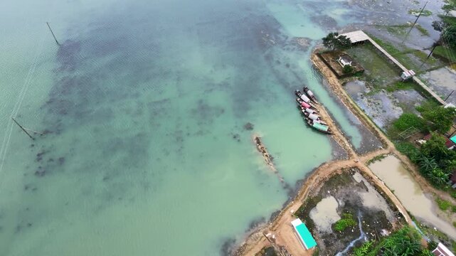 Aerial top-down descending view of turquoise lake shoreline with moored boats, sandy embankment and rural waterfront