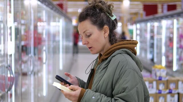 Woman studies smartphone and food package in supermarket aisle, carefully reviewing details with thoughtful expression. Shopper weighs ingredient information while navigating food allergy concerns.