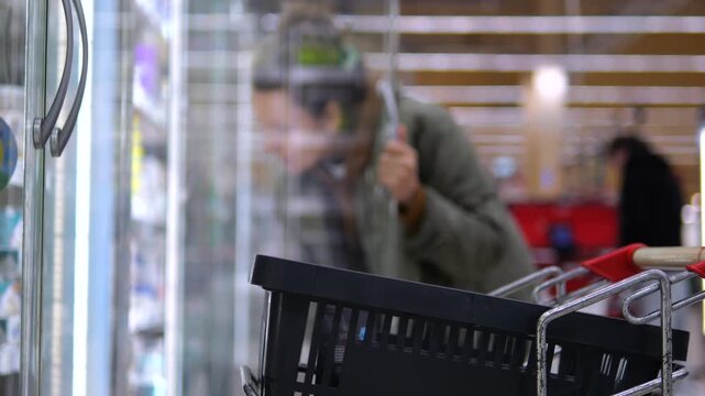 Woman leans into supermarket freezer, reaching for frozen goods behind glass door. Focused shopper searches shelves beside black basket and red cart in bright grocery store, bending to access