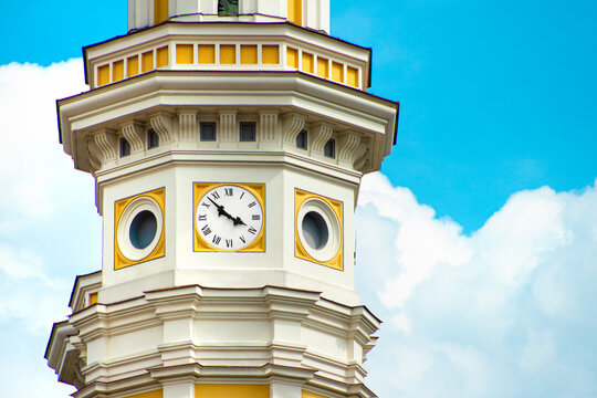 Vintage city clock tower. Old town hall tower. Clock face with Roman numerals. Yellow and white architectural details. European clock tower blue sky. Historic tower against sky. City landmark travel