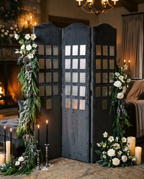 Moody winter wedding seating chart mockup. Dark velvet folding screen with blank metallic cards, pine garlands, white roses, and candles by a cozy fireplace.