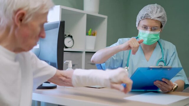 Medical professional examines senior citizen holding records while discussing arm injury diagnosis in clinical setting. Healer checks vulnerable patient condition with serious concern in well-lit room