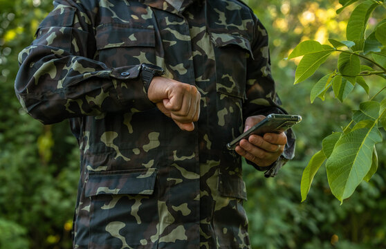 Camouflaged soldier checks smartphone outdoors, glancing at wristwatch while holding phone in one hand. Military personnel consults mobile device among green foliage, with face outside frame.