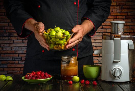 Cook presents bowl of chopped green apples beside juicer, ready for fresh juice preparation. Chef in black jacket holds fruit above counter with raspberries and kitchen ingredients.