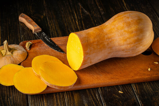 Butternut squash lies on wooden board with knife nearby. Cut end reveals vivid orange flesh, and sliced rounds emphasize fresh kitchen prep in rustic still life styling with cozy, appetizing feel.