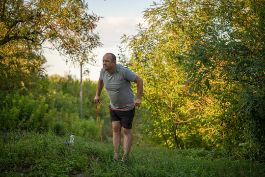 Middle aged man exercises with resistance band on grassy path, leaning forward in focused effort. Outdoor workout takes place among leafy trees, with small water bottle nearby in soft evening light.