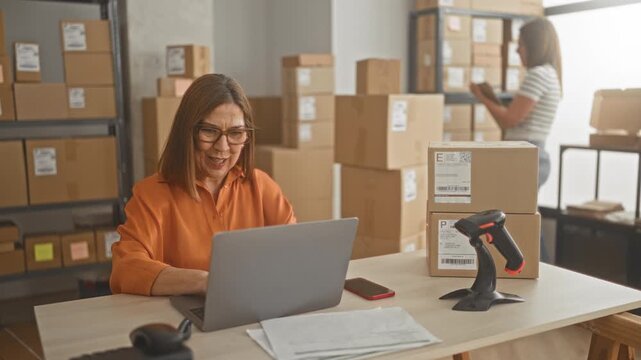 Woman typing on laptop amid stacked packages and barcode scanner in building; entrepreneurial focus productivity.
