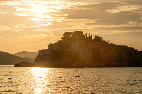Sunset silhouette of rocky island over shimmering sea with distant hills on horizon