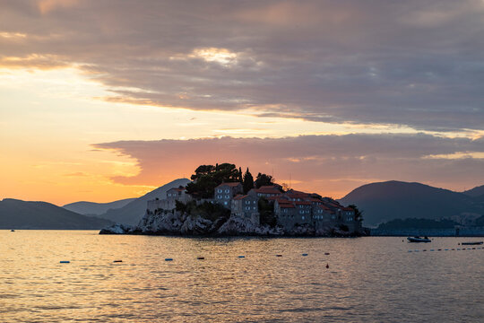 Colorful sunset view of small rocky island with stone buildings, pine trees, and calm sea reflections from coastside panorama with warm golden light, distant mountains, and pastel sky