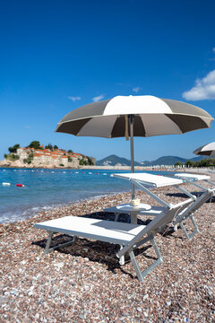 Sunlit beach loungers under striped umbrella on pebbled shore with calm sea and distant coastal town on horizon