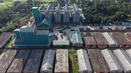 Aerial view of a large industrial grain silo complex with storage warehouses and trucks parked in the facility yard during a sunny day © ridwanhassan