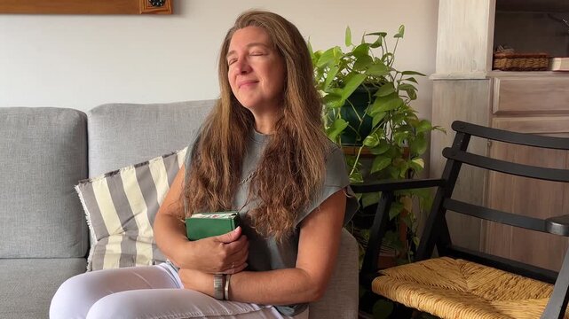 A peaceful and relaxing moment is being enjoyed by a mature woman while sitting on a sofa at home with a book on her hands.