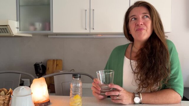 Beautiful woman with long hair taking a relaxing break. She is sitting in her kitchen drinking water, smiling and enjoying the moment