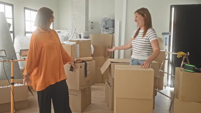 Woman mother and daughter embrace with arm around shoulder, leaning on stacked cardboard moving boxes in building, smiling and surveying the new space; family joy.