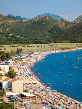 Crowded Mediterranean beach with sunbathers, colorful umbrellas and calm sea, backed by mountains and coastal village along curved shoreline in summer daylight from elevated viewpoint