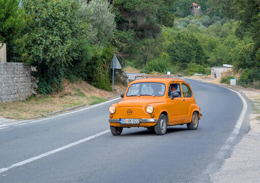 August 25, 2026, Budva, Montenegro, Orange vintage compact car in front view driving along winding curving country road past stone wall and dense green trees in clear daylight with visible asphalt
