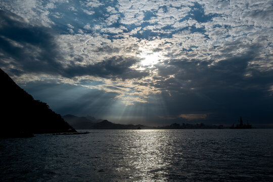 Sunlight pierces through clouds at sunset, illuminating the water surface in Angra dos Reis. Mountains and a distant city skyline are visible along the horizon near the ocean.