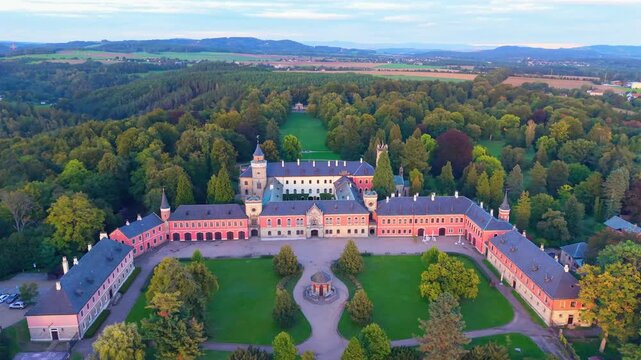 Aerial view of the historic Sychrov Castle featuring its distinctive red facade, sprawling green lawns, and dense surrounding forest in Sychrov, Liberec Region, Czechia.