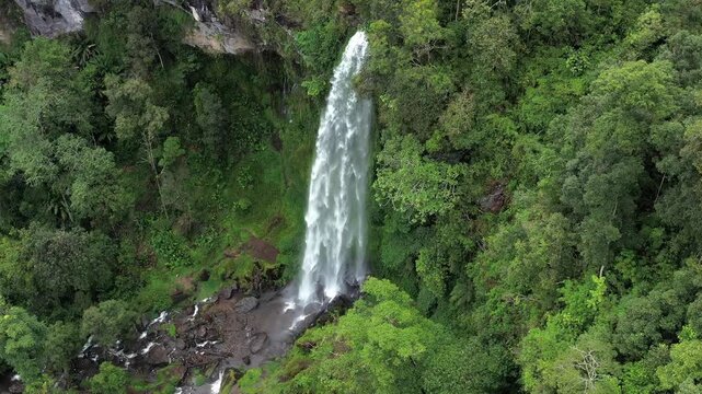 Aerial view of Waterfall Kayu Aro cascading down into a rocky pool surrounded by dense tropical rainforest in Kayu Aro, Jambi, Indonesia.