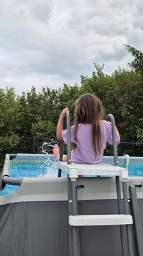 Happy little girl sitting on a ladder of an above-ground swimming pool, splashing water with her feet in a green sunny backyard during summer vacation.