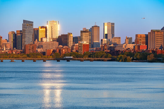 The Back Bay skyline of Boston, United States, rises beyond the Charles River and Harvard Bridge, with sunlit glass towers, brick buildings and riverside trees reflected in the water.
