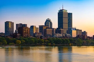 The urban skyline of the Back Bay neighborhood in Boston seen across the Charles River at sunset featuring tall architectural towers, colorful evening reflections, and tree lined riverfront banks. © Jose Luis Stephens