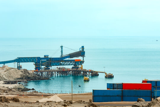 Copper concentrate loading infrastructure at Puerto Punta Padrones, Chile, with a blue conveyor and shiploader extending over a coastal pier beside stacked shipping containers and a rocky shoreline.