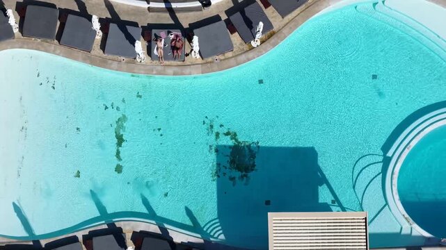 Aerial commercial footage looking directly down over a large outdoor swimming pool in Laganas Zakynthos Greece with turquoise water sun loungers parasols poolside terrace and two people relaxing