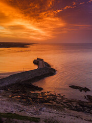 Fototapeta premium Aerial view of coastal pier at golden sunset. Warm light reflects on water and stone breakwater. Calm sea meets land with sandy beach nearby. Spiddal, Ireland, Galway bay. Travel and tourism.