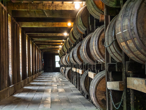 Oak barrels of aging bourbon, stacked on wooden racks along a rick house aisle, with chains, glowing overhead bulbs, and a bright window at the far end in warm, golden light