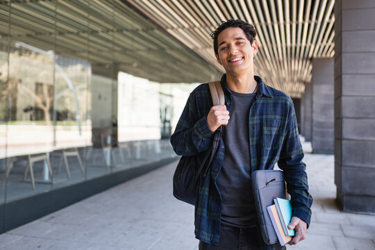 Young latin student carrying laptop books backpack walking outdoors near modern university building