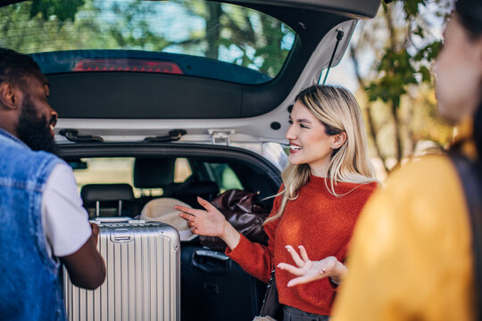 A stylish suitcase sits in the trunk of a car while friends engage in an animated conversation. The scene is set outdoors with trees in the background, capturing a vibrant and cheerful mood as they pr