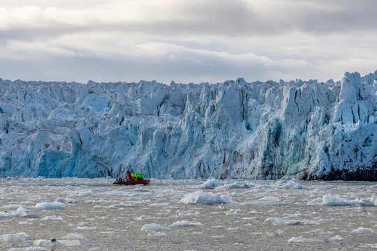 Expedition tourists in an inflatable zodiac boat cruising directly below a towering blue glacier front surrounded by floating ice, Spitsbergen, Svalbard, Norway