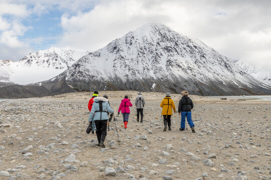 Group of expedition hikers crossing a barren Arctic plain toward massive snow-covered mountains under cloudy summer sky, Spitsbergen, Svalbard, Norway