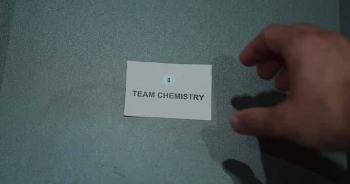 A hand beside a team chemistry paper card on a gray background, representing teamwork, collaboration and workplace cohesion