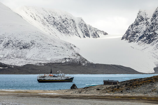 Remote summer Arctic landscape with ship, small cabin, blue fjord water and glacier framed by steep rugged mountains, Spitsbergen, Svalbard, Norway