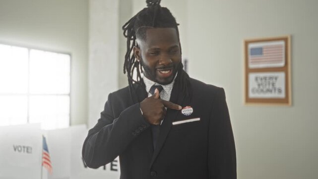 Man in suit pointing finger to 'i voted' sticker on his lapel at a polling station with american flag and vote signs visible; civic pride.