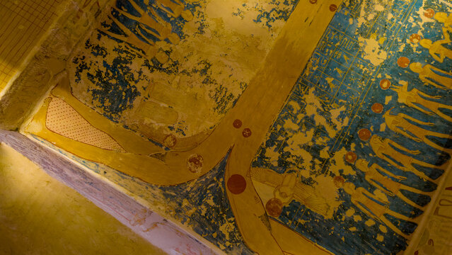 Ancient Egyptian tomb ceiling painting featuring goddess Nut arched over earth with winged scarab and rows of deities depicted in traditional yellow and blue pigments on stone wall surface.