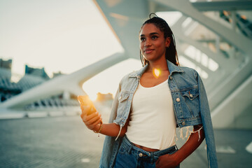 Young woman standing in a modern architectural setting at sunset, holding a smartphone and looking away