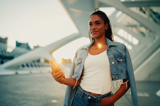 Young woman standing in a modern architectural setting at sunset, holding a smartphone and looking away
