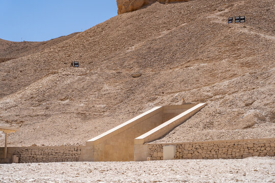 Stone staircase leading down into ancient Egyptian tomb entrance carved into side of dry, rocky desert mountain under clear blue sky in Valley of Kings near Luxor, Egypt.