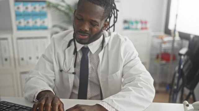 Man doctor in white coat wearing stethoscope rubs neck while typing on computer at desk in medical building with file shelves and cart visible; fatigue.