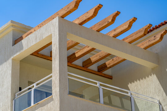 Low angle view of modern building exterior featuring textured beige stucco wall and wooden pergola structure shading balcony with glass railing under clear blue sky on sunny day.
