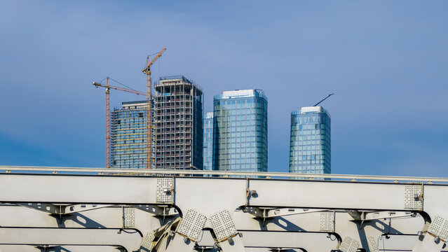 Modern skyscrapers under construction with towering cranes against blue sky behind metal bridge structure.