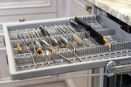 Dirty cutlery basket pulled out from modern dishwasher in kitchen, filled with unwashed spoons, forks, knives, and kitchen utensils covered in food residue.