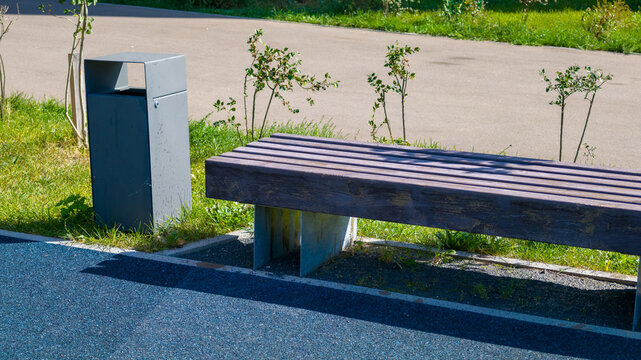 Wooden park bench with metal frame sits next to grey trash bin on sunny day in public park with paved path and green grass in background.